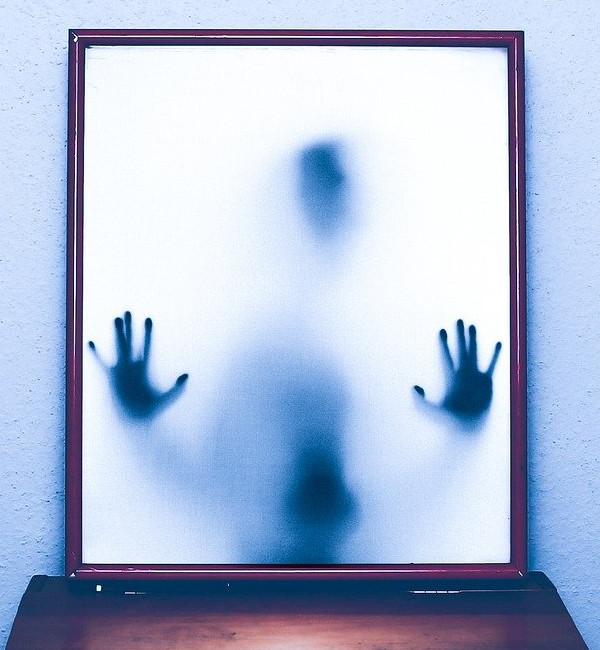 Un espejo cuadrado con fondo blanco en el que una figura difuminada apoya las manos desde el interior